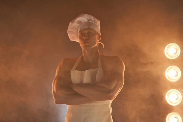 Портрет мускулистого шеф-повара в белом фартуке и шляпе шеф-повара, позирующего со сложенными руками на дымчатом фоне домохозяйки-мужчины. Муж на кухне. Жестокий мясник. - Фото, изображение