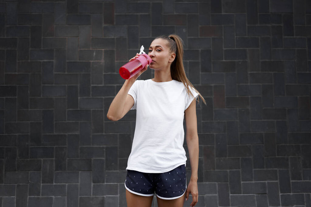 Yorgun kadın sabah antrenmanından sonra kırmızı şişe su içer. Genç atletik kadın sağlıklı yaşam tarzı kavramının ardından şehir sokaklarının kara duvarında duruyor.  - Fotoğraf, Görsel