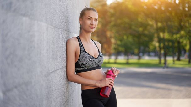 Tired Woman drink water red bottle after morning workout Young athletic female rear view standing leaning wall city street park background after jogging Healthy lifestyle concept Copy space - Foto, imagen