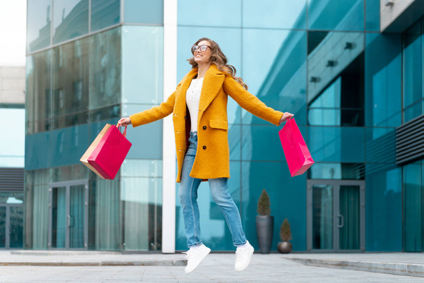Business Woman With Shopping Bags Dressed Yellow Coat Jumping With Happiness Outdoors Corporative Building Caucasian Female Business Person Near Office Building Stylish Businesswoman Season Sale - Фото, зображення