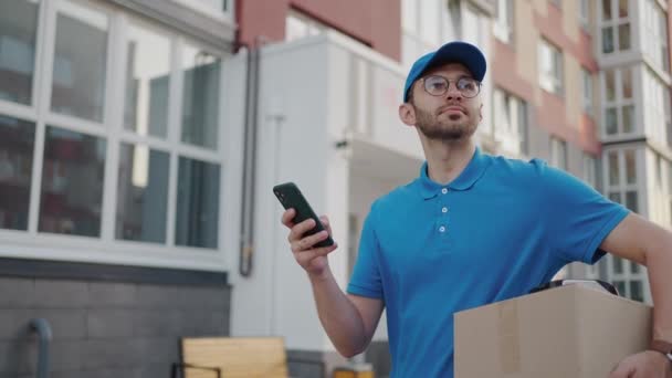 白人の若いかなりの男は、青色のキャップで配達労働者の通りを歩いて、ルートを探して、スマートフォンを使用している間、カートンボックスを運ぶ。男性宅配便で小包のタップと電話でテキストメッセージ. - 映像、動画
