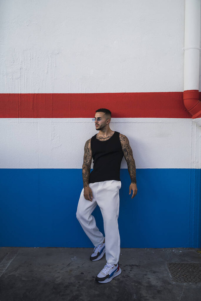A hot caucasian man with arm tattoos in a black t-shirt and white pants posing near a colorful wall - Photo, Image