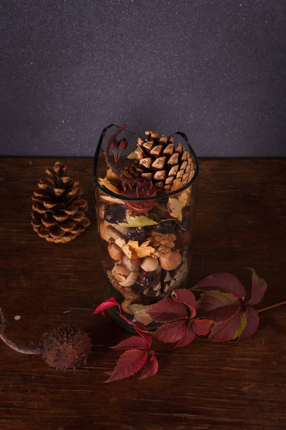 Transparent glass vase with pine, fir cones, nuts, chestnuts, dry leaves, acorns, hip rose berries on dark background. Autumn composition, vertical shot - Photo, Image