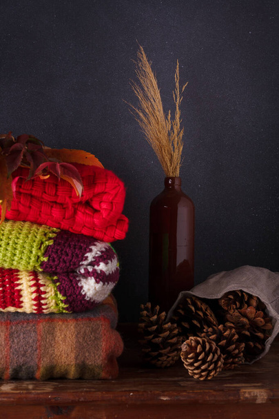 Plaids tricotés lumineux, chandails, vase marron avec feuilles sèches et cônes de pin sur fond gris profond. Composition d'automne, concept hygge - Photo, image
