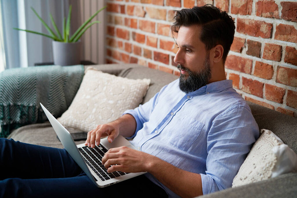 Чоловік у сорочці сидить на дивані і використовує ноутбук
                                - Фото, зображення
