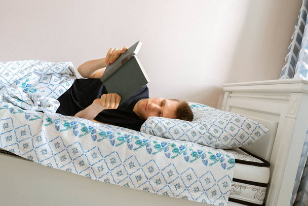 a young male lying in the bed and read a book in the evening before going to sleep at home bedroom - Photo, image