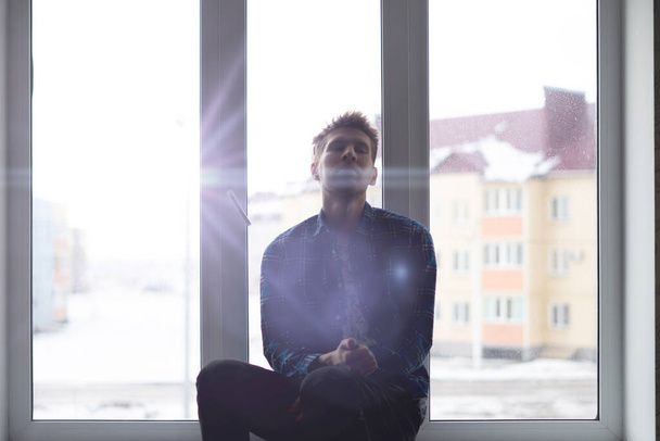 una triste y solitaria silueta de hombre pensativo sentado en un alféizar de ventana y mirando al aire libre lejos, - Foto, imagen