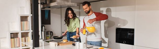 Man pouring orange juice near smiling girlfriend with pancakes in kitchen, banner  - Photo, Image