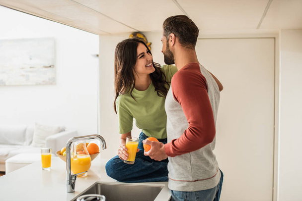 Smiling woman looking at boyfriend with orange near juice in kitchen  - Fotografie, Obrázek