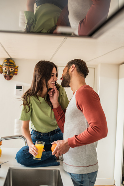 Cheerful man touching chin of girlfriend with glass of orange juice in kitchen  - Photo, image