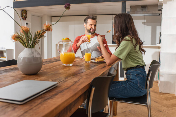 Cheerful man with orange juice talking to girlfriend near pancakes and laptop on table  - 写真・画像
