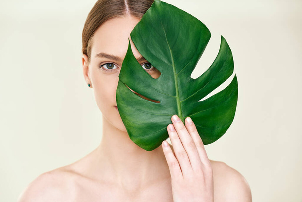 Close up portrait of young beautiful woman with perfect smooth skin covering her eye with monstera tropical leaf - Foto, Imagem