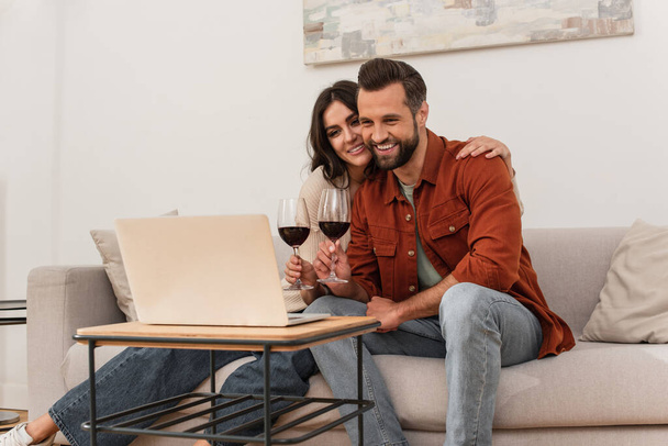 Smiling woman embracing boyfriend with glass of wine near laptop at home  - Foto, afbeelding