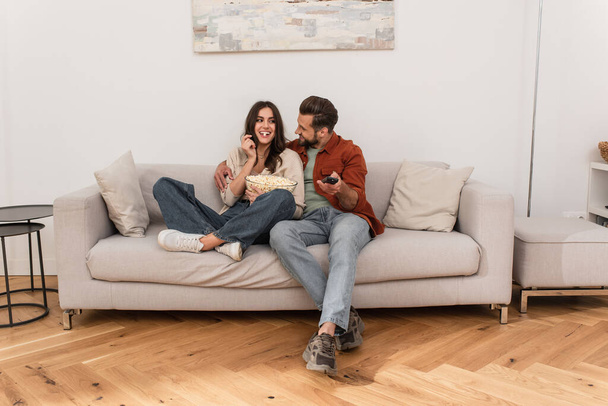 Homme souriant avec télécommande regardant petite amie manger du pop-corn à la maison  - Photo, image