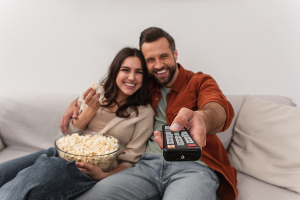 Remote controller in hand of blurred man near cheerful girlfriend with popcorn  - Фото, изображение