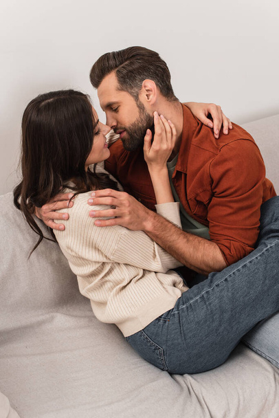 Vista lateral do jovem casal beijando e abraçando no sofá  - Foto, Imagem