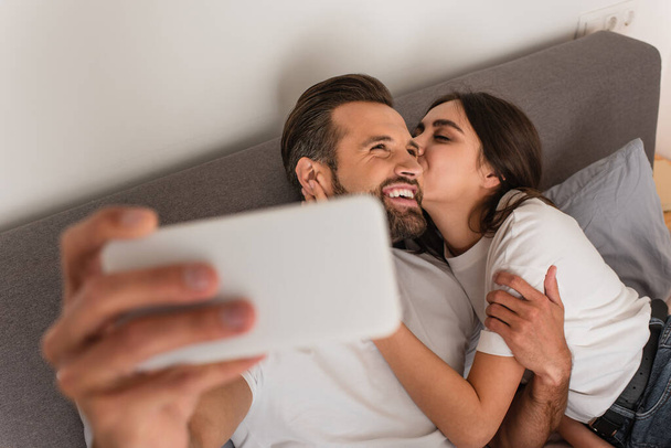 Woman kissing boyfriend during selfie on blurred smartphone on bed  - Fotó, kép