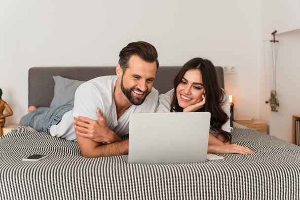 若いカップルはベッドの上の携帯電話の近くのノートパソコンを見て  - 写真・画像