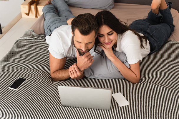 Smiling woman looking at laptop near boyfriend and cellphone on bed  - Φωτογραφία, εικόνα