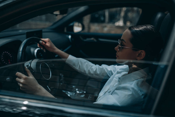 Piękna młoda kobieta siedzi za kierownicą w czarnym nowoczesnym samochodzie. Widok przez otwarte okno. - Zdjęcie, obraz