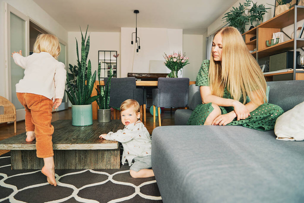 Giovane madre che riposa sul divano mentre i bambini piccoli giocano e saltano in soggiorno - Foto, immagini