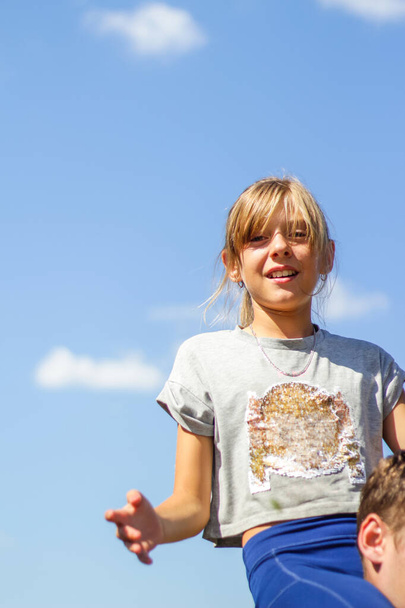 Ξανθιά κοπέλα που χαμογελάει στο καλοκαιρινό φόντο του μπλε ουρανού. Κάθετα. Να κάνει χειρονομίες στους ώμους των αντρών. Δωρεάν ευτυχισμένη παιδική ηλικία. Εκτός εστίασης. - Φωτογραφία, εικόνα