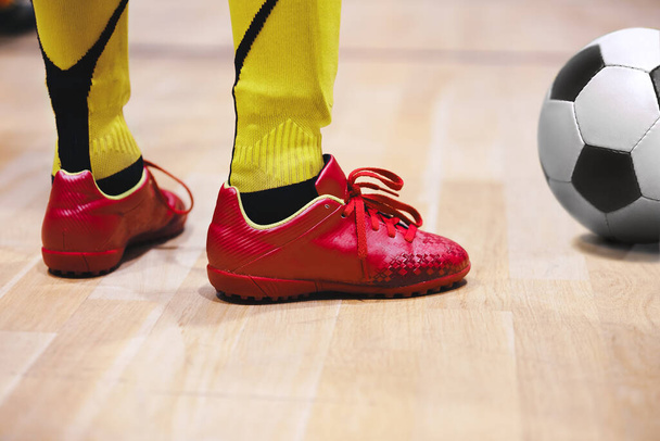 School children playing indoor soccer at physical education class. Legs of young boy standing in red cleats on wooden floot with soccer ball - Photo, Image