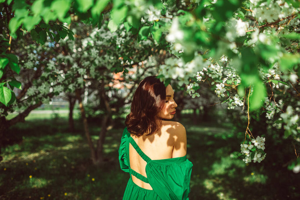 A young beautiful girl stands in the park under a blossoming apple tree, wearing a beautiful bright green dress. - Fotoğraf, Görsel