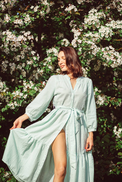 Genç ve güzel bir kız parkta çimlerin üzerinde çiçek açan beyaz bir elma ağacının altında oturuyor, açık renk bir elbise giyiyor.. - Fotoğraf, Görsel