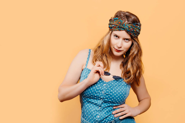 Young blonde girl with a bright headscarf looking at the camera holding sunglasses with a denim top on a yellow background. summer concept. copy space - Fotoğraf, Görsel