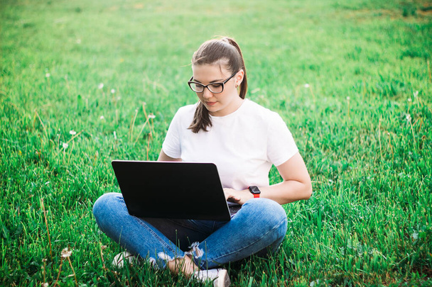 νεαρή όμορφη κοπέλα που εργάζονται σε ένα φορητό υπολογιστή σε εξωτερικούς χώρους, κάθεται στο γρασίδι. - Φωτογραφία, εικόνα