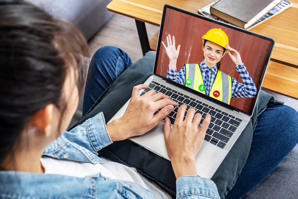 保護ヘルメットの女性ビルダーは、クライアントにノートパソコンの画面から手を振って。自宅滞在中の修理コンセプトに関するリモートコンサルティング - 写真・画像