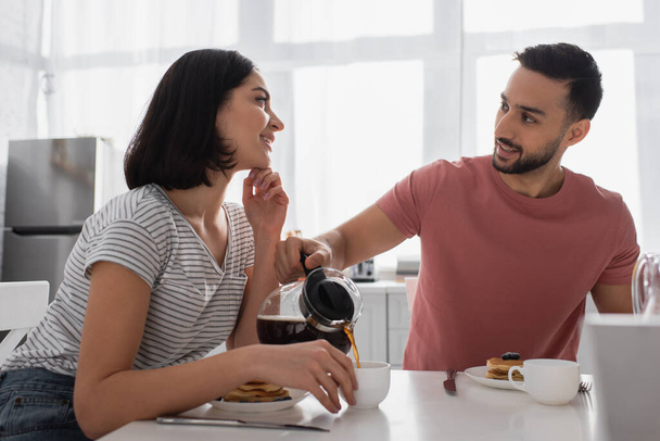 sorrindo jovem com a mão perto do rosto olhando para o namorado derramando café em xícara na cozinha - Foto, Imagem