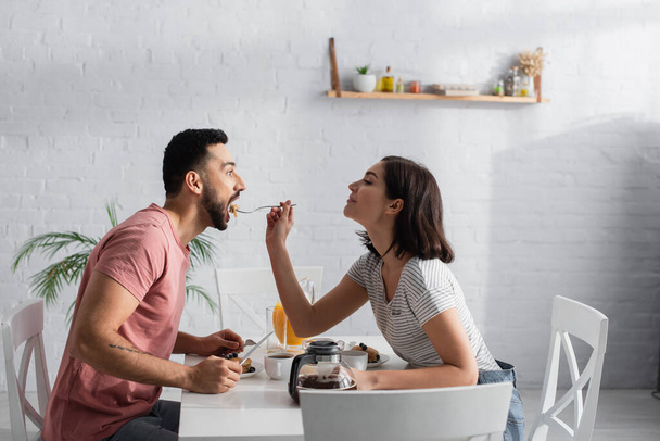 smiling young woman feeding boyfriend with pieces of pancakes on fork in kitchen - Photo, Image
