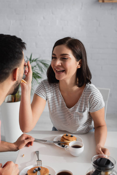 positive young woman feeding boyfriend with pieces of pancakes on fork in kitchen - Foto, imagen