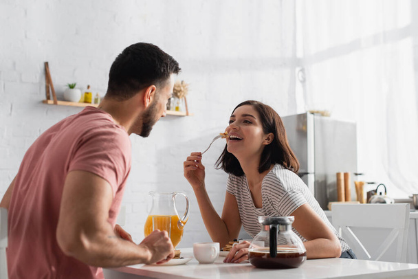 mujer joven positiva comiendo panqueques cerca de novio en la cocina - Foto, Imagen