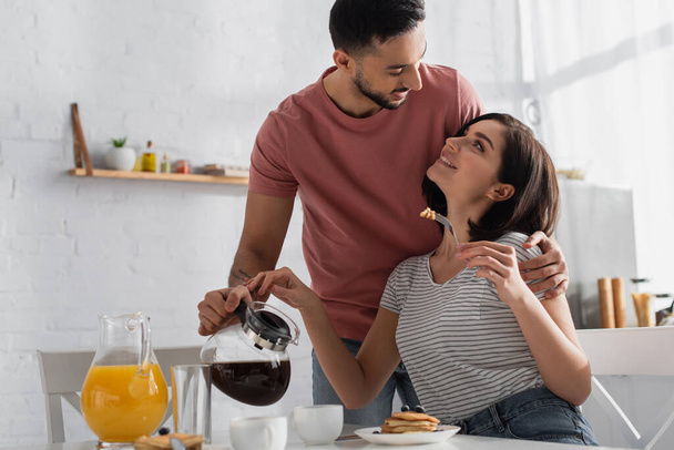happy young man hugging girlfriend with pieces of pancakes on fork and pouring coffee from pot to cup in kitchen - Photo, Image