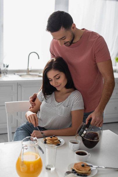 smiling young man hugging girlfriend eating pancakes and pouring coffee from pot to cup in kitchen - Photo, image