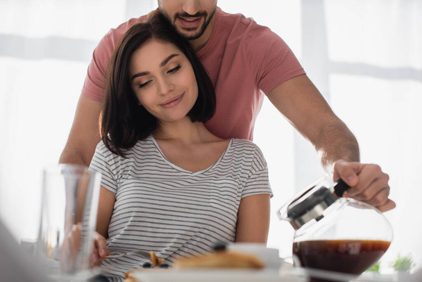 young man hugging girlfriend with breakfast and pouring coffee from pot to cup in kitchen - Photo, Image