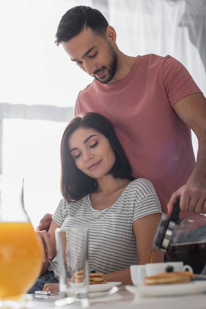 smiling young man hugging girlfriend with breakfast and pouring coffee from pot to cup in kitchen - 写真・画像