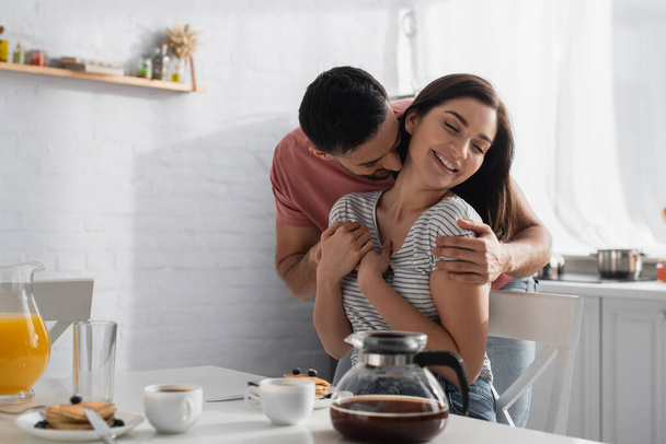 happy young couple hugging and kissing near table with breakfast, coffee and orange juice in kitchen - Foto, afbeelding