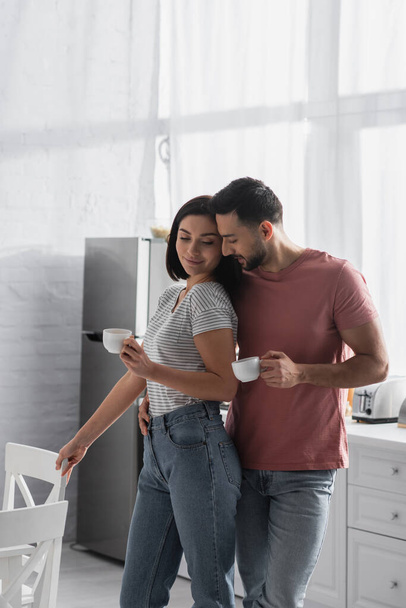 happy young couple hugging with coffee cups near chairs in kitchen - Foto, Bild