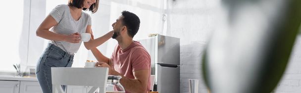 pareja joven tocándose suavemente y sosteniendo tazas con café en la cocina, pancarta - Foto, Imagen