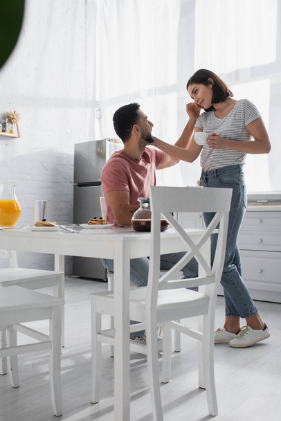 若いカップルがキッチンで朝食とコーヒーでテーブルの近くに優しく接触し - 写真・画像