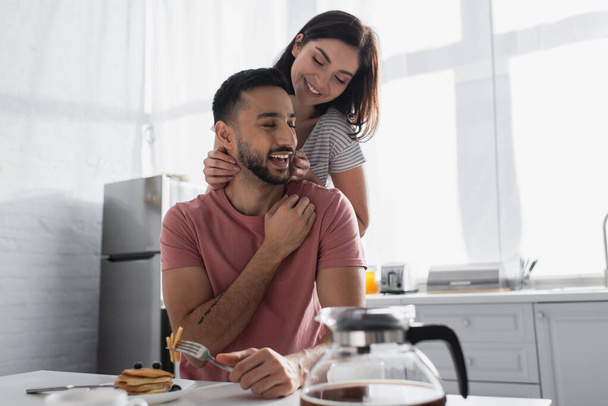 smiling young woman gently touching boyfriend near table with breakfast and coffee in kitchen - Photo, Image