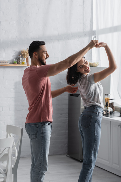 positive young couple in casual clothes dancing with holding hands in modern kitchen - Photo, Image