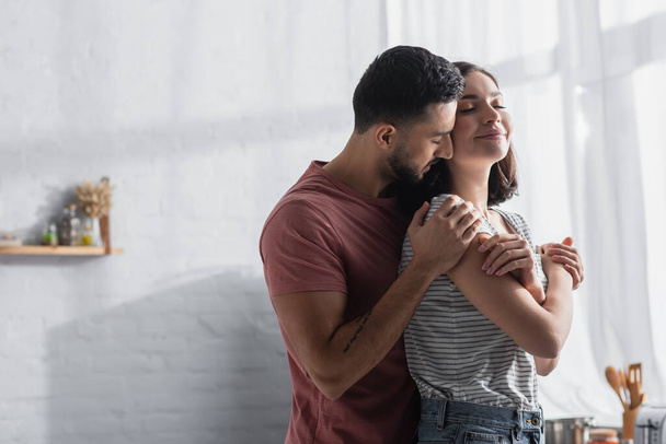 smiling young couple with closed eyes hugging in modern kitchen - Foto, afbeelding