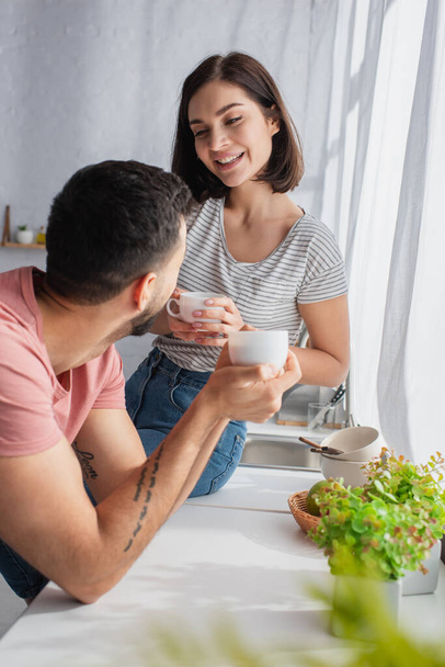 positive young couple holding white cups with coffee near window in kitchen - Foto, immagini