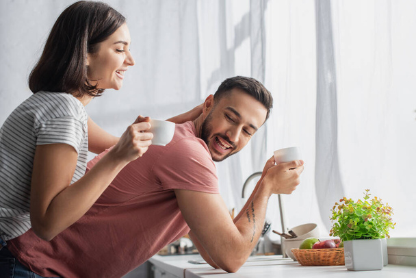 hymyilevä nuori nainen tilalla valkoinen kuppi kahvia ja koskettaa poikaystävä lähellä ikkunaa keittiössä - Valokuva, kuva