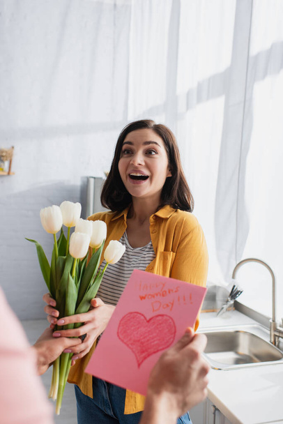 young man presenting bouquet of flowers and greeting card to excited girlfriend in kitchen - Photo, Image
